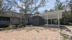 Modern two-story house with gray siding, a large carport, and a brick driveway surrounded by trees and landscaped plants under a partly cloudy sky.