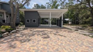 Two-car carport with a modern, flat roof design, adjacent to a dark gray house. The driveway is paved with tan and brown bricks. Trees and greenery surround the property.