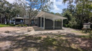 Modern gray house with a flat roof carport, surrounded by trees and a paved driveway, on a sunny day.