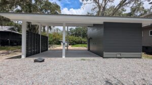 A modern carport with a flat roof and adjacent storage unit, set on a gravel driveway, with trees and grass in the background.