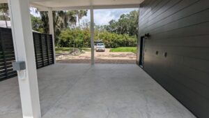 Covered concrete carport with black paneled wall, open sides, and a white vehicle parked on a brick driveway outside, surrounded by trees and greenery.