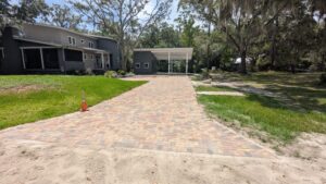 A brick driveway leads to a carport next to a modern gray house, with a single orange traffic cone on the left side and trees surrounding the area.