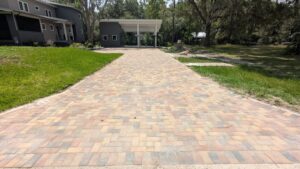 A paved brick driveway leads to a covered carport next to a gray house, surrounded by grass and trees under a clear sky.