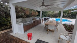 Covered outdoor kitchen and dining area with a grill, cabinets, wicker chairs, and a wooden table; pool and greenery in the background.