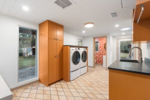 Modern laundry room with orange cabinets, front-loading washer and dryer, tiled floor, countertop, sink, and large window with garden view.