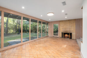 A spacious room with large floor-to-ceiling windows, a tile floor, a brick fireplace with a fire burning, and a door leading to a wooded backyard.