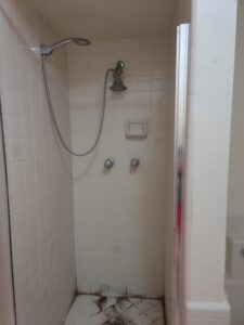Shower with white tile walls, a handheld showerhead, two knobs, a soap dish, and visible stains on the floor.