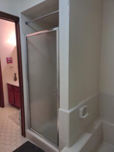 A bathroom with a glass shower door, white tiles, a soap holder, and a partial view into another room with a vanity and hexagonal floor tiles.