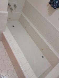 White tiled bathtub with jets, hexagonal floor tiles, wall-mounted faucets, and a small tiled ledge along the tub's edge. A section of patterned fabric is visible on the upper right.