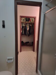 View from a bathroom showing a walk-in closet with clothes hanging and shoes on the floor. Shower and toilet paper holder are also visible.