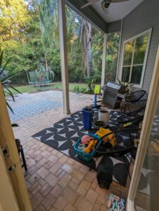 Covered patio with outdoor seating, scattered toys, a grill, and chalk. Backyard has trees, pavers, and a trampoline.