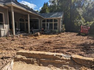 A backyard with disturbed soil and tire tracks, construction equipment, and workers near a house, indicating ongoing landscaping or renovation work.