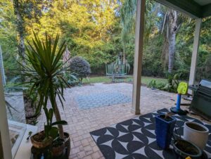 A covered patio with potted plants overlooks a brick backyard, trampoline, kids’ baseball toy, and surrounding trees in the background.