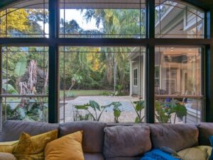 A gray sofa with yellow pillows is in front of large windows, showing a view of a patio with plants, trees, and part of a house exterior in the background.
