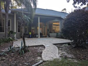 Back view of a house with a paved patio, arched doorway, open sliding door, small dog, scattered items, and surrounding plants and trees.