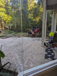 A small excavator is digging in a backyard near a patio with pavers, viewed from inside a house through a glass door.