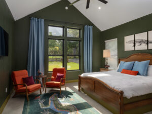 Bedroom with green walls, wood bed, orange chairs, blue curtains, large window, crocodile artwork, and a lizard rug on the floor.