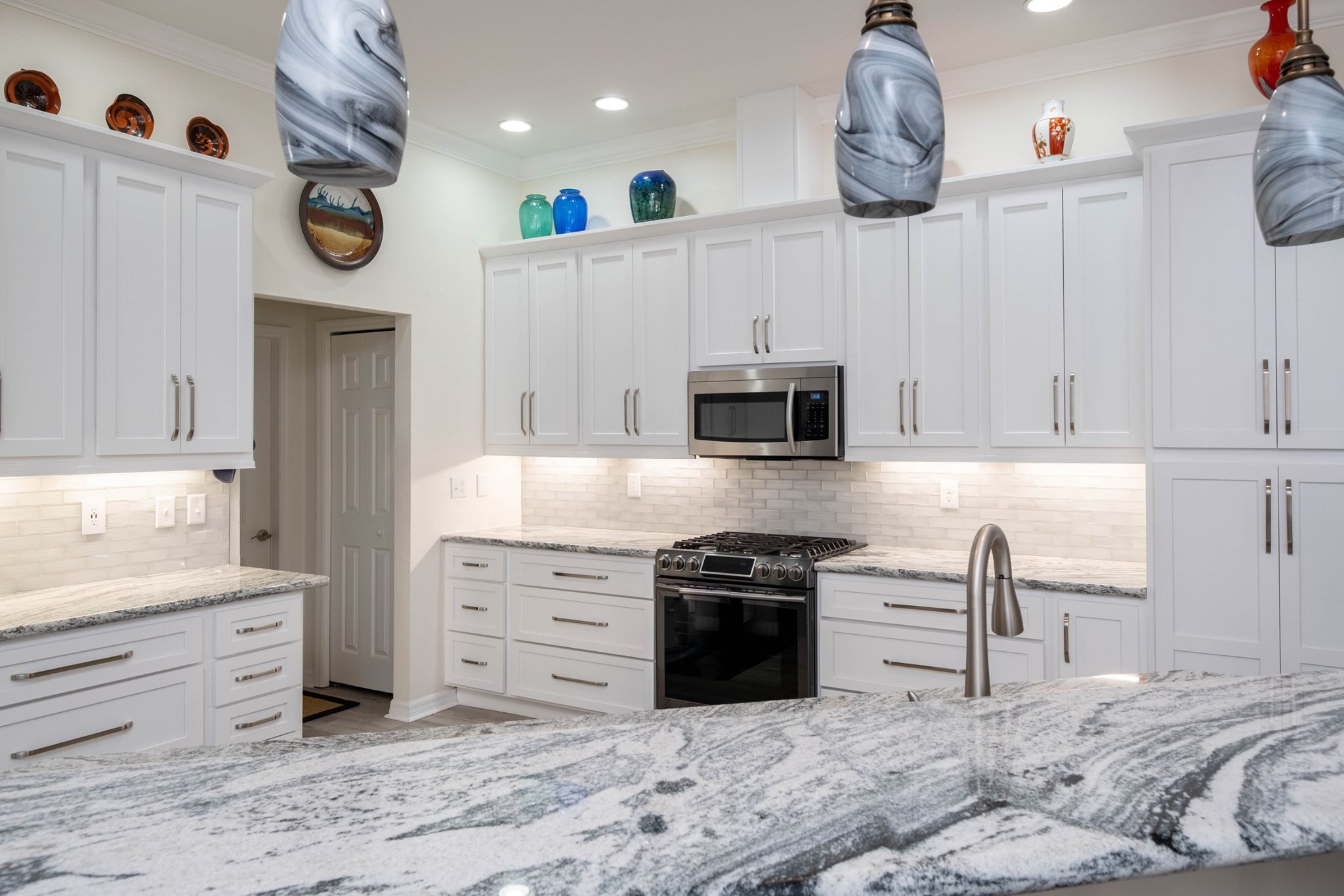 Modern kitchen with white cabinets, stainless steel appliances, marble countertops, gray and white pendant lights, and colorful glass vases displayed above the cabinets.