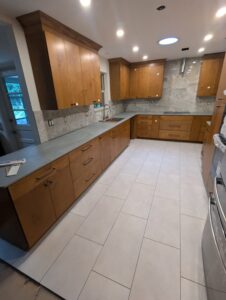 Modern kitchen with light wood cabinets, gray countertops, tiled backsplash, and white floor tiles; appliances and fixtures are not fully installed.