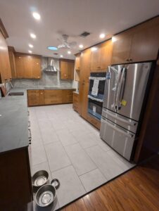 Modern kitchen with wooden cabinets, stainless steel appliances, tile floor, and three pet bowls on the ground near the entrance. Some appliances still have protective packaging.