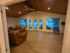 A living room with wooden floors and ceiling, brown leather sofa, large windows, and glass doors overlooking a lake at dusk.