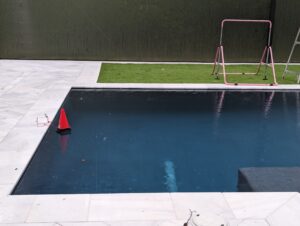 A red safety cone floats in a backyard pool near the edge; a small soccer goal and artificial grass are in the background.
