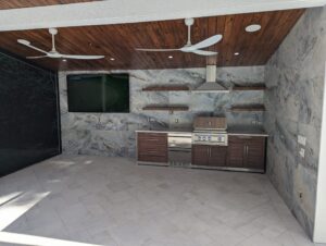 Modern outdoor kitchen with stainless steel appliances, wood cabinets, mounted TV, stone walls, floating shelves, two ceiling fans, and a wood-paneled ceiling.