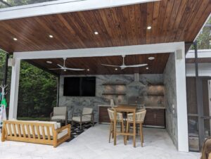Covered outdoor kitchen and lounge area with wooden ceiling, ceiling fans, TV, bar seating, and patio furniture.