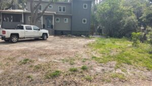 A white pickup truck is parked on a dirt driveway beside a gray two-story house surrounded by trees and greenery.