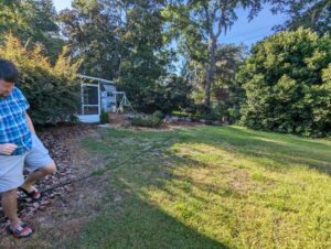 A man in sandals walks on grass near a garden with a shed, swing set, and trees in the background on a sunny day.